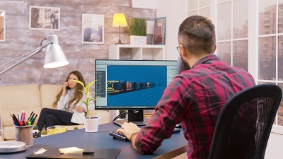 Over the shoulder shot of young game developer working from home while his girlfriend walks in the backgorund